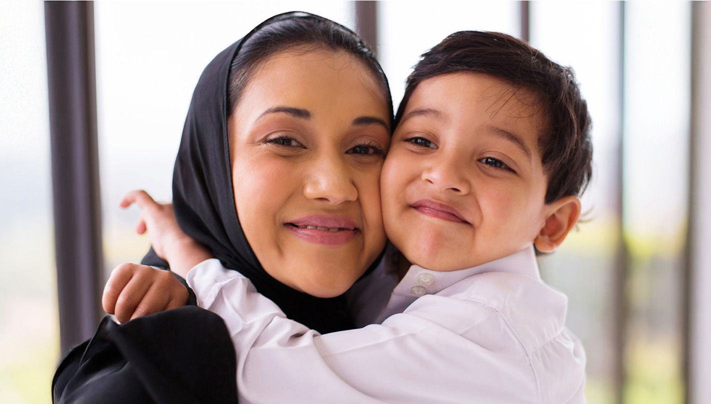 Woman wearing a hijab and dress with a young boy hugging her around her neck.
