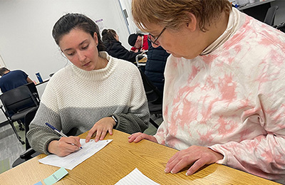 Image of a young woman writing on paper as another woman looks on.