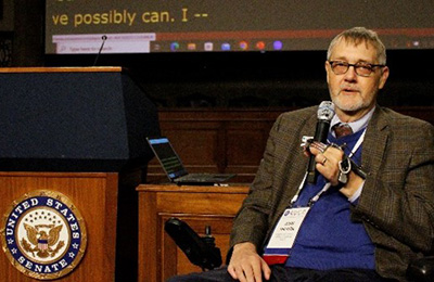 John Tschida, a smiling man with glasses, graying brown hair and goatee, wearing a tweed suit, white shirt, and blue patterned tie, holding a microphone.