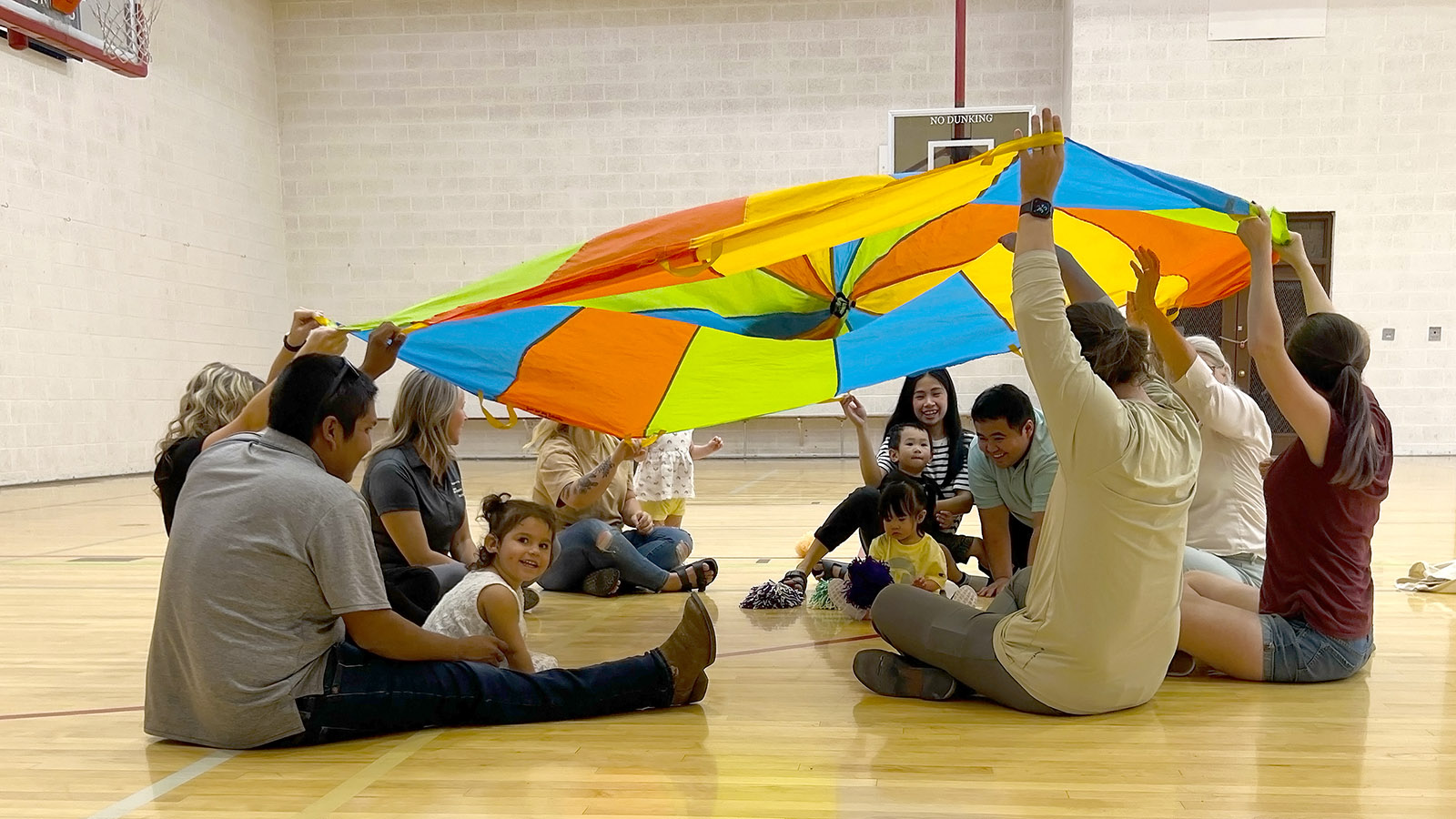 A group of adults and children play under a large, multicolored parachute