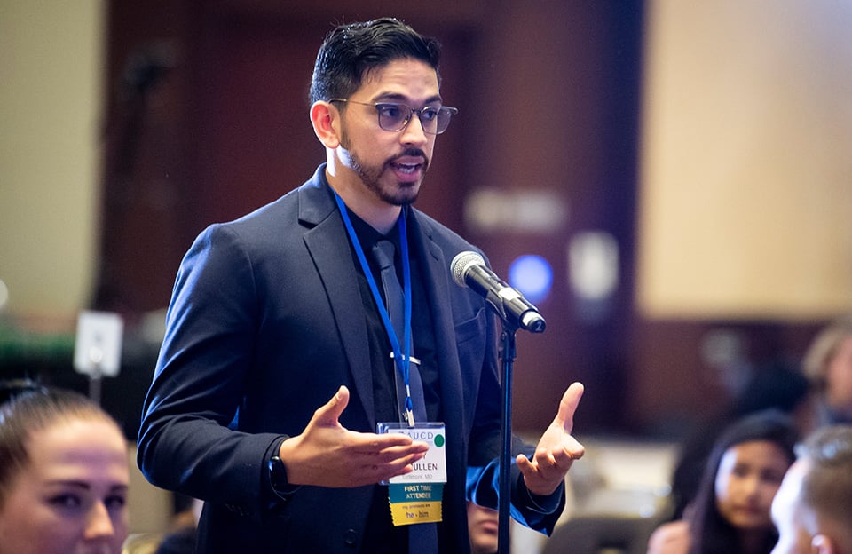 Image of a young man with facial hair and glasses asking a question in a microphone.