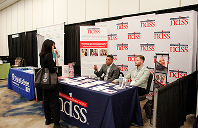 Image of the NDSS booth with one Conference attendee speaking to two booth attendants.