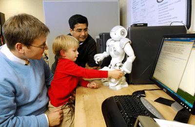 A child is sitting on a man's lap, playing with a robot on a desk while a man watches on.Two 