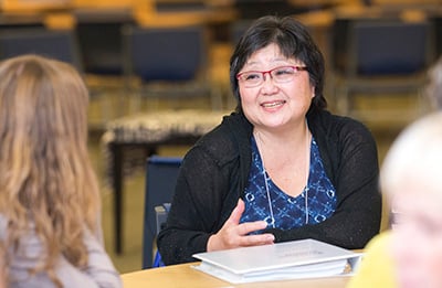 Asian woman with short black hair and glasses sitting at a table talking to another woman.