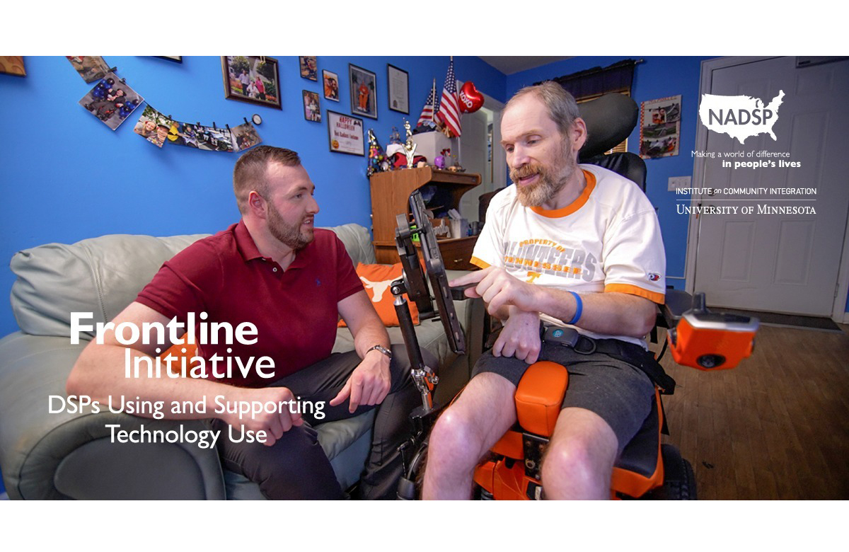 A man sits at home, typing on a screen attached to his wheelchair. His direct support professional looks at him from the sofa.