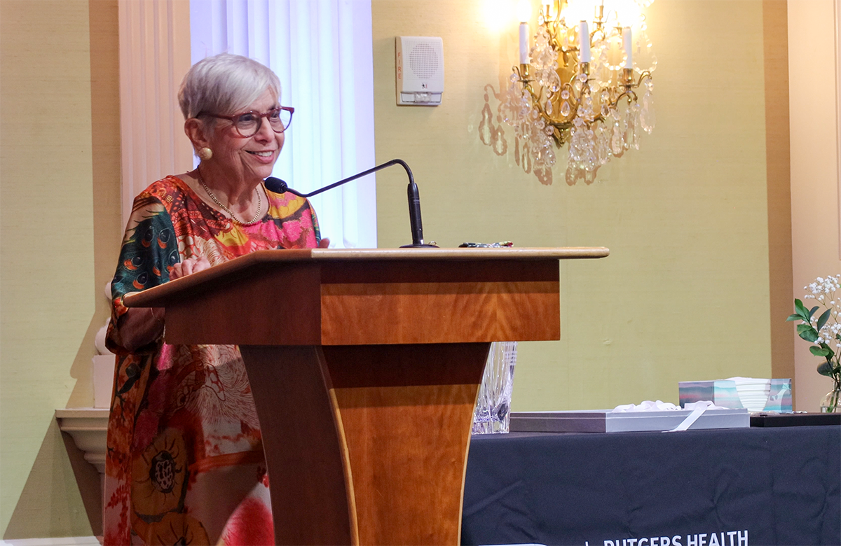 Deborah M. Spitalnik, a white woman with short grey hair, is wearing a dress and standing at a podium. 