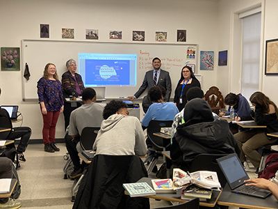 GUCEDD employees and teachers stand in front of a classroom filled with students. The slide being projected onto the board reads, "What makes you feel like you belong in the classroom?"