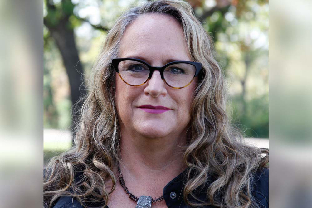 Amy Hewitt, a white woman with long brown hair wearing coral glasses and a blouse.