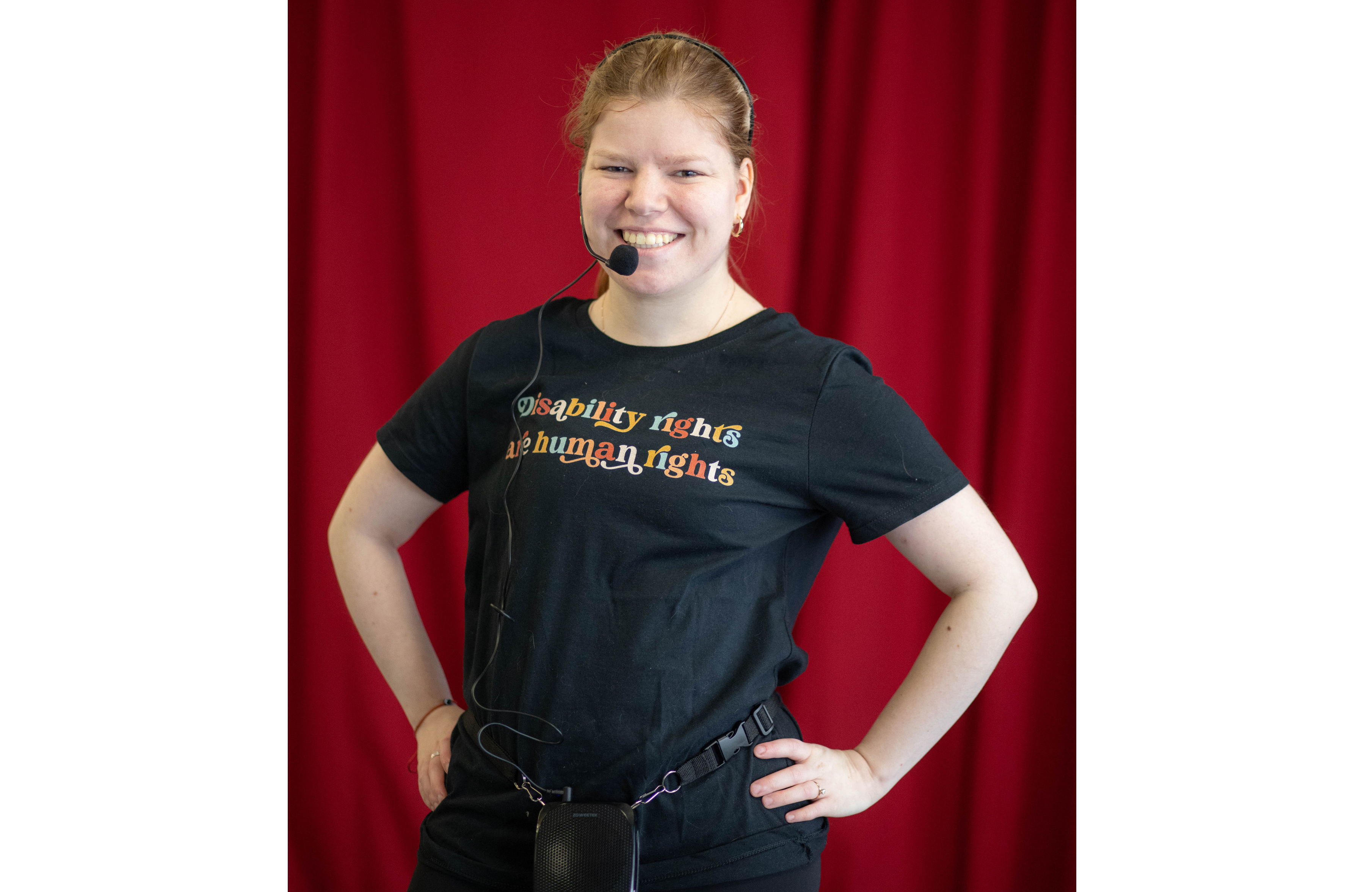 Sophie Iverson is wearing a headset and a t-shirt that reads, "Disability rights are human rights." She is smiling. There is a red curtain behind her.