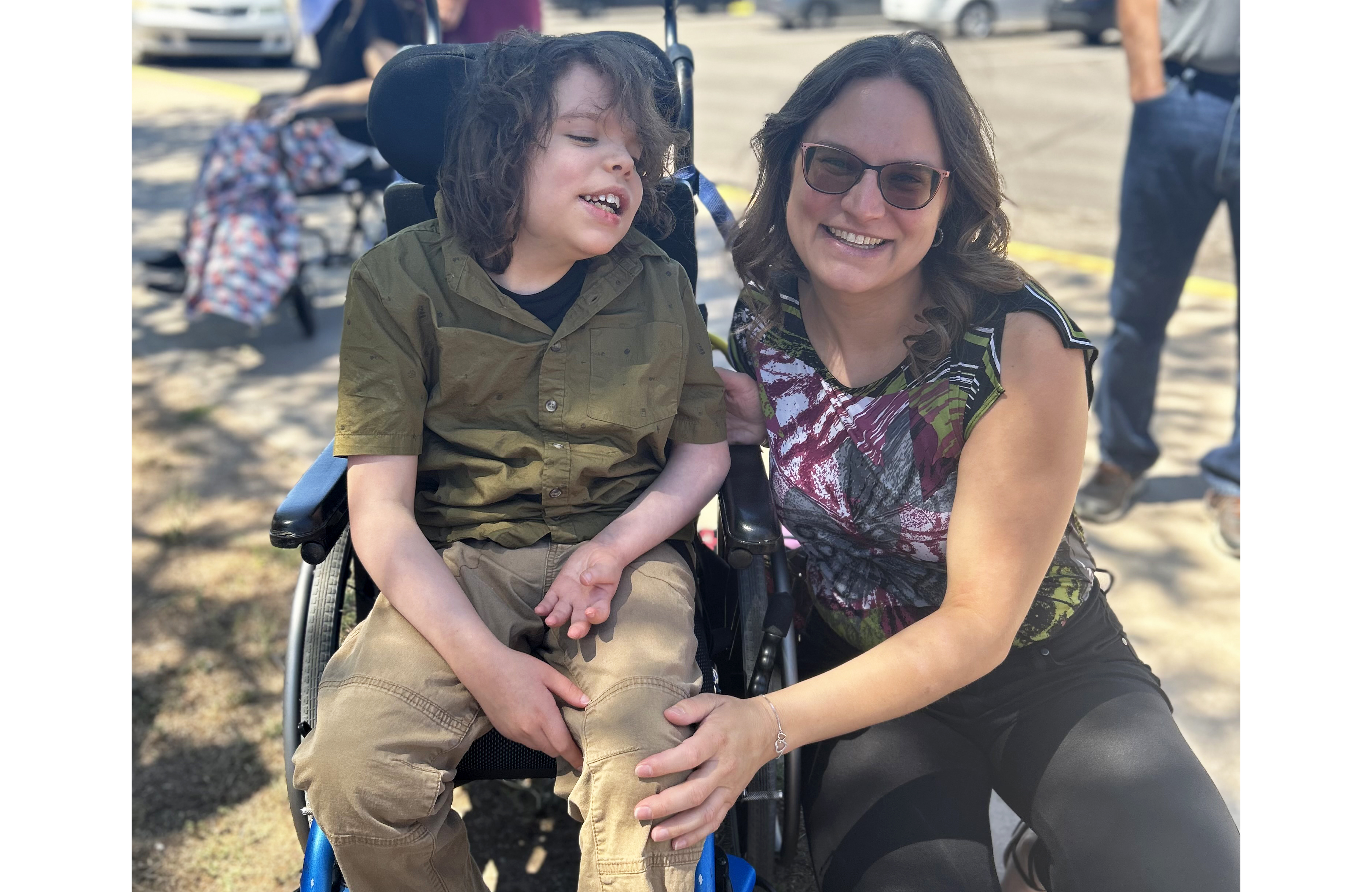 Jaclyn Larson and her son Aidan outside. Aidan is sitting in a wheelchair, and Jaclyn is kneeling next to him.