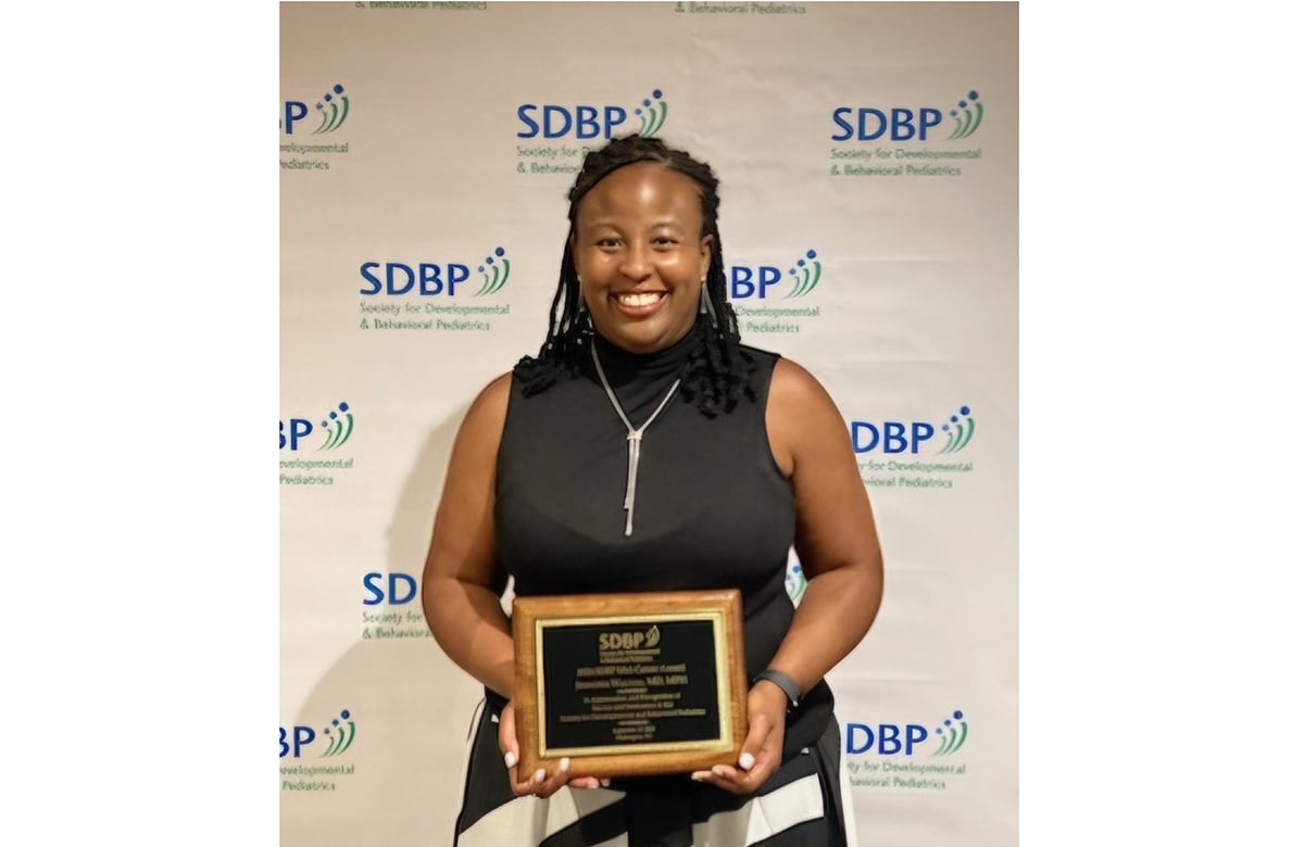 Dr. Walton, a black woman wearing a dress and holding an award in front of a banner.