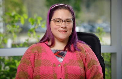 A white woman with long purple hair and glasses wearing a blouse and sweater and using a power wheelchair.