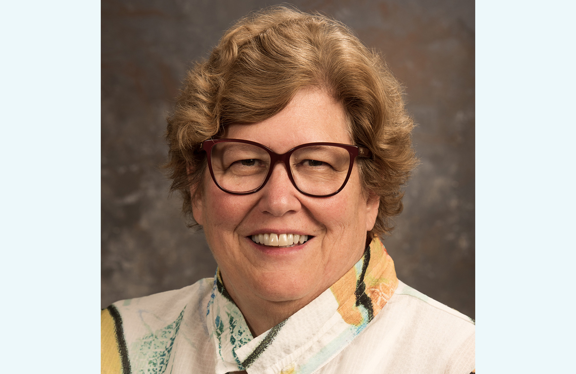 This is a headshot of Sandy Root-Elledge, a smiling older white woman with short brown hair. She is wearing glasses and a blazer.