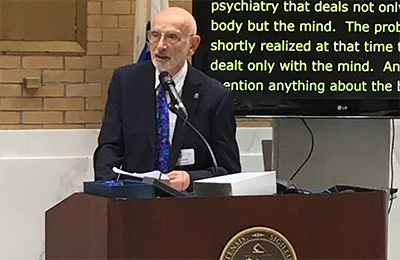 Imaage of  Dr. Ludwik Stefan Szymans, an older white man in a suit, standing at a podium.