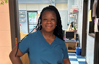 A young Black female Next Steps student smiles and poses for a picture in front of the door to the daycare nursery where she is interning. She has long braids and is wearing a short-sleeved V-neck scrubs top. 