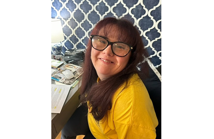 Headshot of Alexandra "Allie" Hayes. Allies is wearing a yellow shirt and glasses while smiling into the camera. In front of her is a table covered with various documents, a computer mouse, and a lamp.