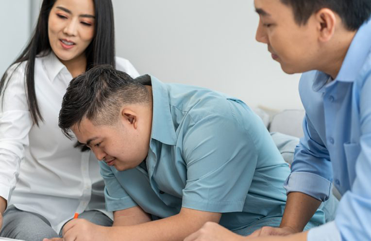 An adult brother and sister sit with their adult brother with Down syndrome as he signs a document.