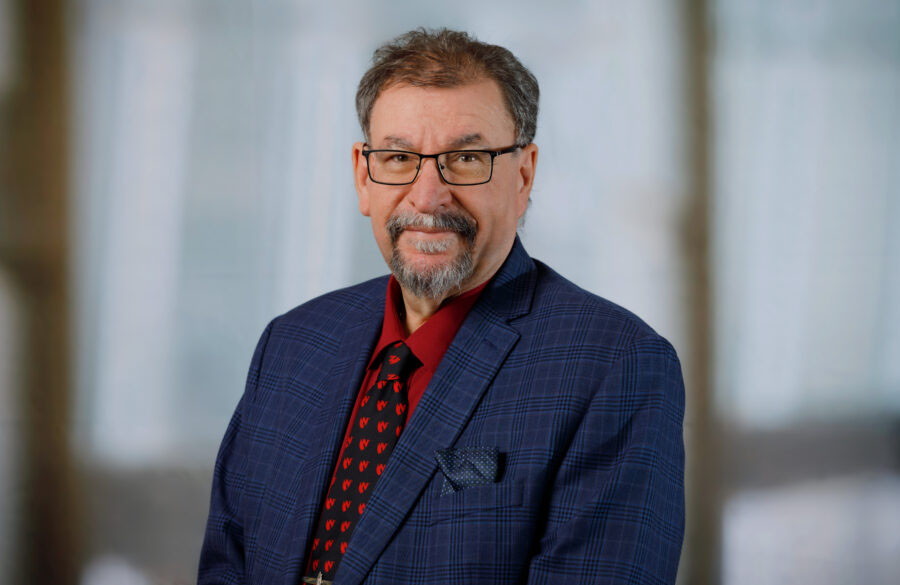 An older white man with grayish hair and beard is wearing glasses, a jacket, a shirt, and a tie. 