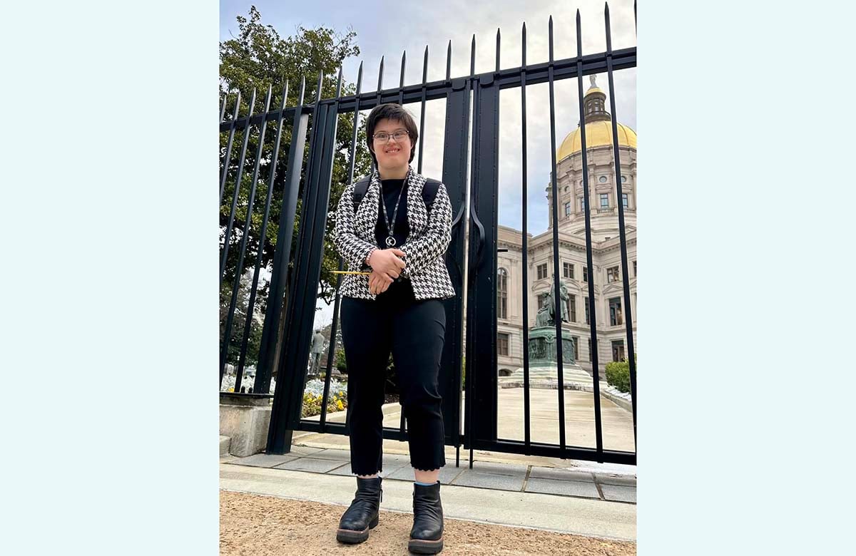 Image of Martha, a white woman with short black hair wearing black pants and hats with a checkered blazer standing with her hands crossed in front of Georgia state capitol. 