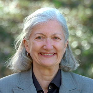A headshot of Elise McMillan, a smiling white woman with gray shoulder-length hair, blue eyes, small gold hoop earrings, a black button-down blouse, and a gray blazer.