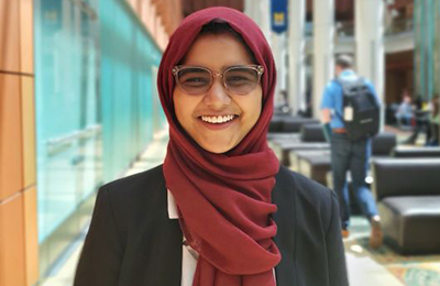 A headshot of Tazia Miah. She is wearing a dark scarf, glasses, and a jacket, smiling in an outdoor setting. 