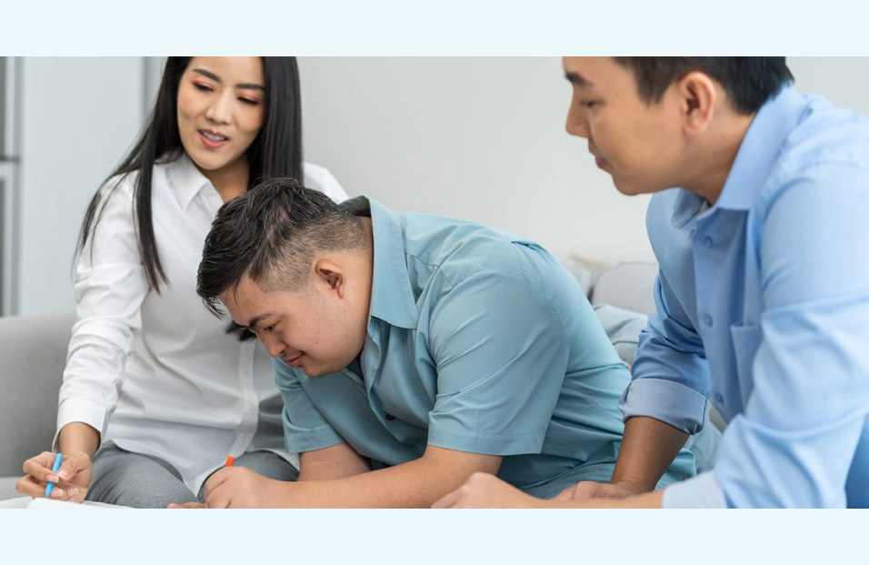 A young woman and a young man, possibly a sister and brother, flank another young man, this one with Down syndrome, as he fills out a document in front of him.