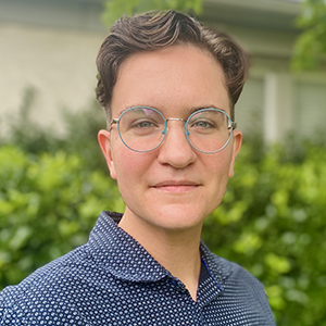 Headshot photo of Gale Hann, a smiling transmasculine nonbinary person who has light olive skin and short brown hair. They are photographed in front of a green, leafy background wearing glasses and a navy-blue shirt.