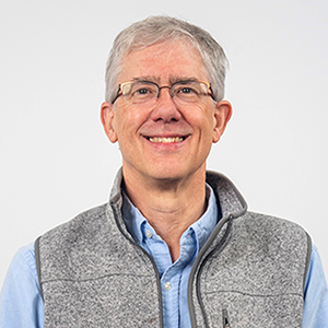 Image of John Butterworth, a white man with short grey hair wearing glasses, he is wearing a shirt and vest.